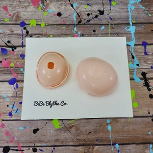 May include: Two pink plastic doll head molds, one with a hole in the center and the other with a textured surface. The molds are on a white surface with the text "BeBe Blythe Co."