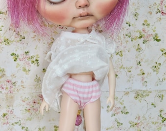 May include: A doll with pink hair wearing a white dress and pink and white striped underwear. The doll has a beaded necklace and is standing in front of a floral patterned background.