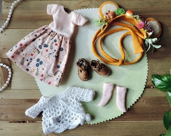 Könnte beinhalten: Ein Set Puppenkleidung bestehend aus einem rosa Blumenkleid, einem weißen Häkelcardigan, einem Paar rosa Socken, einem Paar braunen Schuhen und einem Blumenhaarband.