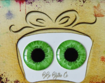 Puede incluir: Un par de ojos de vidrio verde con pupilas negras, sobre un fondo blanco. Los ojos están enmarcados por un recorte de madera de una caja de regalo con un lazo. El fondo es una superficie texturizada amarilla con salpicaduras de pintura.