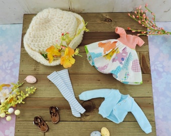 Könnte beinhalten: Ein Puppen-Outfit-Set mit einem weißen Häkelhut, einem rosa-weißen Kleid mit Gingham-Muster, einem blauen Cardigan und gestreiften Strümpfen. Das Set enthält auch ein Paar braune Schuhe und ein paar Ostereier.