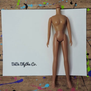 May include: A nude-colored plastic doll torso with no head, arms, or legs. The doll is standing on a white piece of paper with the text "BeBe Blythe Co." written in black.