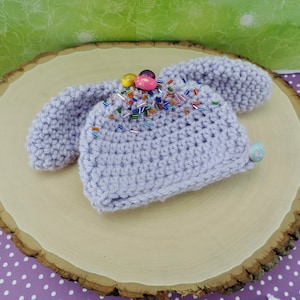May include: A lavender crocheted baby hat with bunny ears and colorful sprinkles on top.