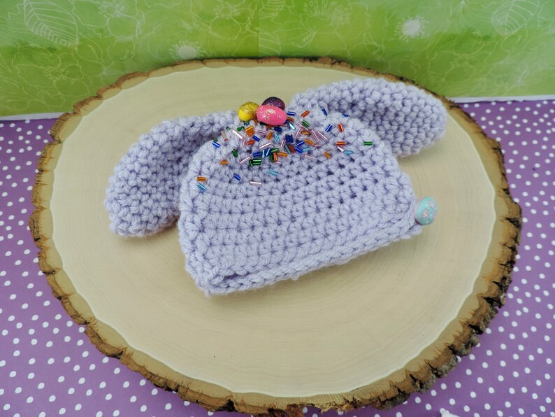 May include: A lavender crocheted baby hat with bunny ears and colorful sprinkles on top.