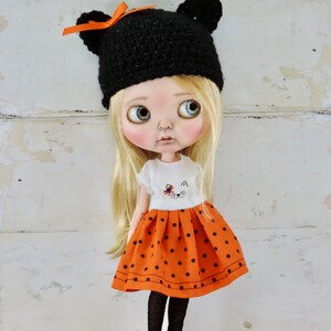 May include: A doll with blonde hair wearing a black crocheted hat with orange ribbon, a white shirt with a cat design, and an orange and black polka dot skirt.