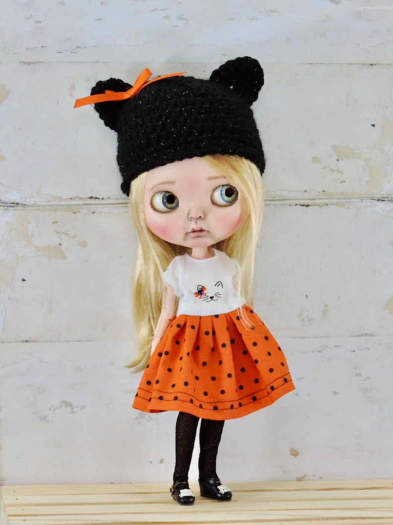 May include: A doll with blonde hair wearing a black crocheted hat with orange ribbon, a white shirt with a cat design, and an orange and black polka dot skirt.
