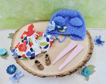 Könnte beinhalten: Ein Set Puppenkleidung bestehend aus einem rot-weißen Kleid mit Erdbeeren und Blaubeeren, einer blauen Strickmütze mit Blumenverzierungen, rosa-weißen gestreiften Strumpfhosen und braunen Schuhen.