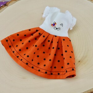 May include: A white doll dress with a black cat face embroidered on the bodice. The dress has a short-sleeved bodice and a full orange skirt with black polka dots.