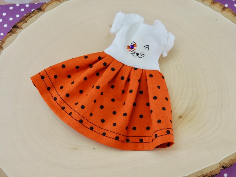 May include: A white doll dress with a black cat face embroidered on the bodice. The dress has a short-sleeved bodice and a full orange skirt with black polka dots.