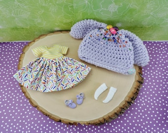 Könnte beinhalten: Ein Set Puppenkleidung bestehend aus einer lila gehäkelten Mütze mit bunten Streuseln, einem gelben und weißen Kleid mit bunten Streuseln und einem Paar weißen Socken.