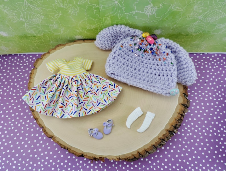 May include: A set of doll clothes including a purple crocheted hat with colorful sprinkles, a yellow and white dress with colorful sprinkles, and a pair of white socks.