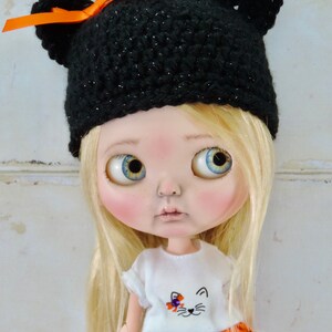 May include: A doll with blonde hair wearing a black crocheted hat with orange ribbon and ear flaps, a white shirt with a cat design, and an orange and black polka dot skirt.