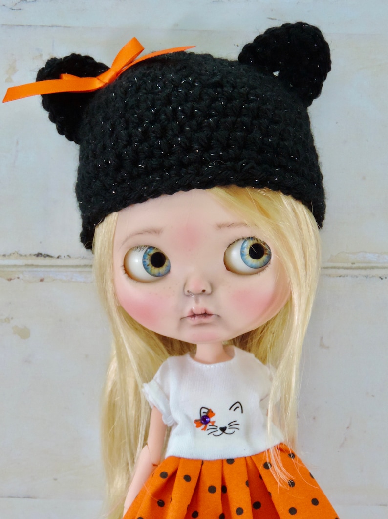 May include: A doll with blonde hair wearing a black crocheted hat with orange ribbon and ear flaps, a white shirt with a cat design, and an orange and black polka dot skirt.