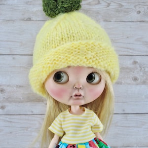 May include: A doll wearing a yellow knitted hat with a green pom-pom, a yellow and white striped shirt, and a colorful skirt with fruit and cherry designs.