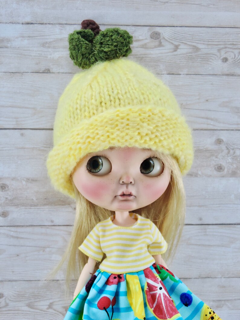 May include: A doll wearing a yellow knitted hat with a green pom-pom, a yellow and white striped shirt, and a colorful skirt with fruit and cherry designs.