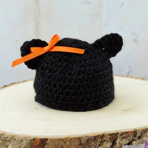 May include: Black crocheted baby hat with sparkly yarn and orange ribbon bow. The hat has two small crocheted ears on top.