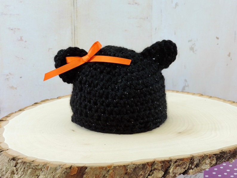 May include: Black crocheted baby hat with sparkly yarn and orange ribbon bow. The hat has two small crocheted ears on top.