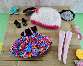 Könnte beinhalten: Ein Puppen-Outfit-Set mit einer weißen Häkelmütze mit schwarzen Ohren, einem schwarz-weißen Blumenkleid mit rosa Trägern, rosa-weißen gestreiften Strumpfhosen und schwarzen Schuhen.