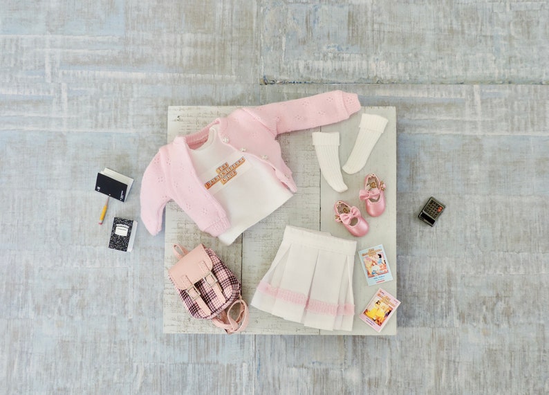 May include: A pink knitted cardigan, a white t-shirt with the text "I'm a little bit of a mess, but I'm a whole lot of awesome", a white pleated skirt with pink trim, a pink plaid backpack, a pair of white socks, pink ballet shoes, two books, a pencil, and a calculator.