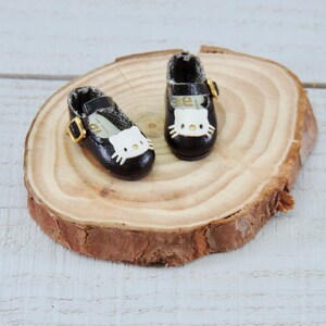 May include: A pair of black doll shoes with a white Hello Kitty face design and gold buckles. The shoes are sitting on a wooden surface.