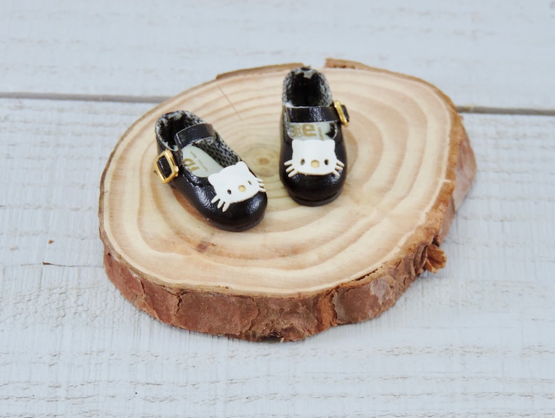 May include: A pair of black doll shoes with a white Hello Kitty face design and gold buckles. The shoes are sitting on a wooden surface.