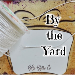 May include: A spool of white cord with the text "By the Yard" and "B&B Blythe Co." on a white card.