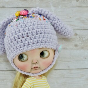 May include: A lavender crocheted bunny hat with colorful sprinkles and candy eggs on top. The hat has a chin strap and is designed for a doll.