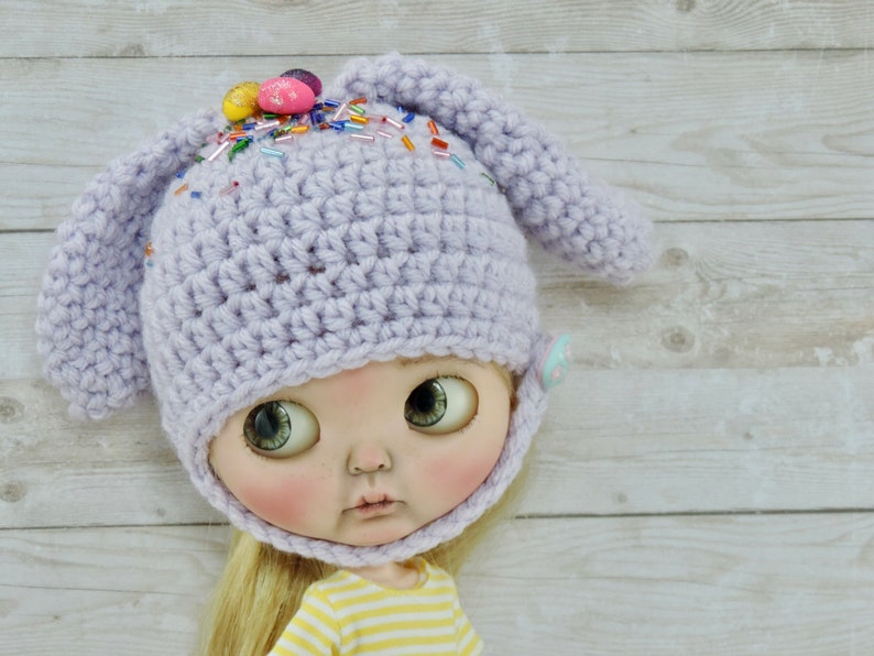 May include: A lavender crocheted bunny hat with colorful sprinkles and candy eggs on top. The hat has a chin strap and is designed for a doll.