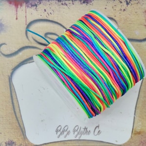 May include: A spool of colorful rainbow cord, featuring shades of red, orange, yellow, green, and blue. The cord is neatly wound on a white base, with the text "Babe Blythe Co" visible. The background has a painted, artistic design.