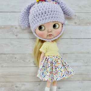 May include: A doll wearing a lavender crocheted bunny hat with colorful sprinkles, a yellow and white striped shirt, and a white skirt with colorful sprinkles. The doll is standing on a light purple wooden platform.