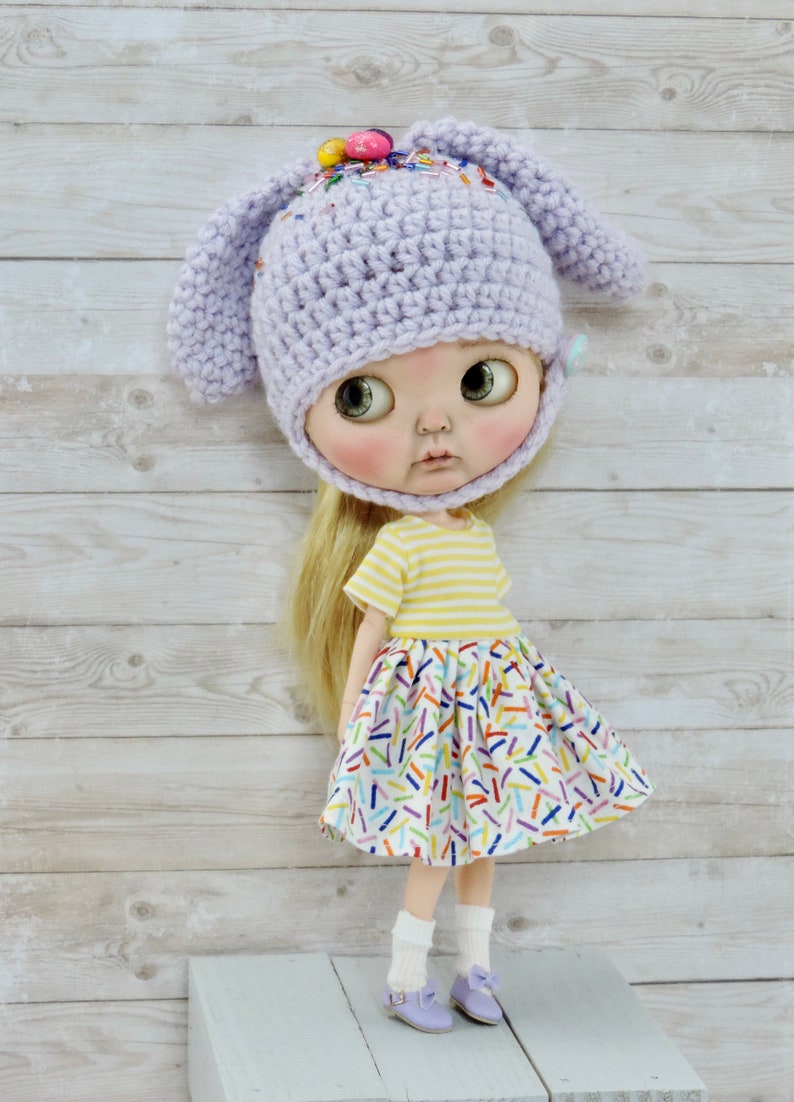 May include: A doll wearing a lavender crocheted bunny hat with colorful sprinkles, a yellow and white striped shirt, and a white skirt with colorful sprinkles. The doll is standing on a light purple wooden platform.