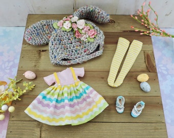 Könnte beinhalten: Ein Puppen-Outfit-Set mit einer grauen und weißen gehäkelten Hasenmütze mit Blumenverzierungen, einem rosa und weißen gestreiften Kleid, gelben und weißen gestreiften Strumpfhosen und blauen Schuhen.