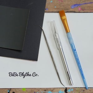 May include: A collection of art supplies including a black paper, a white paper with "BeBe Blythe Co." printed on it, a silver craft knife, a metal tool, and a blue paintbrush with a gold brush head.