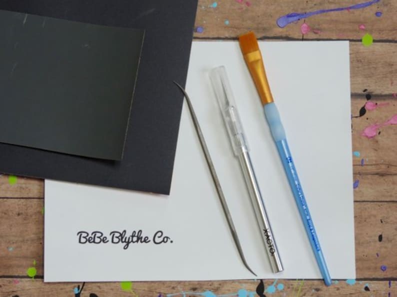 May include: A collection of art supplies including a black paper, a white paper with "BeBe Blythe Co." printed on it, a silver craft knife, a metal tool, and a blue paintbrush with a gold brush head.