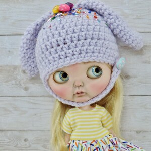 May include: A purple crocheted bunny hat with colorful sprinkles and candy eggs on top. The hat is worn by a doll with blonde hair and a yellow and white striped shirt. The doll is wearing a white dress with colorful sprinkles.