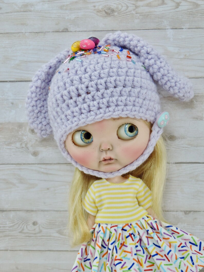 May include: A purple crocheted bunny hat with colorful sprinkles and candy eggs on top. The hat is worn by a doll with blonde hair and a yellow and white striped shirt. The doll is wearing a white dress with colorful sprinkles.