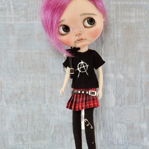May include: A doll with pink hair, wearing a black t-shirt with a white anarchy symbol, a red and black plaid skirt, black tights, and red shoes.