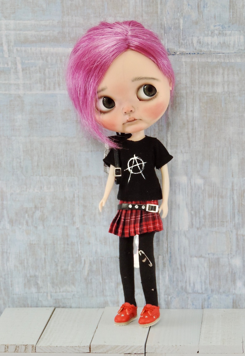 May include: A doll with pink hair, wearing a black t-shirt with a white anarchy symbol, a red and black plaid skirt, black tights, and red shoes.