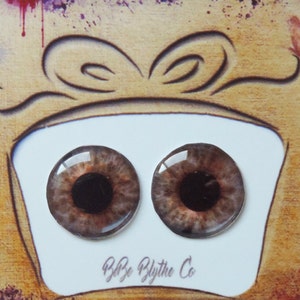 Könnte beinhalten: Zwei braune und schwarze Puppenaugen mit einer hellbraunen Iris und einer schwarzen Pupille. Die Augen werden auf einer weißen Karte mit dem Text "B&B Blythe Co." unten angezeigt. Die Karte befindet sich auf einem braunen Hintergrund mit bunten Farbflecken.