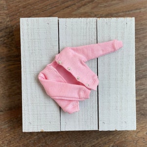 May include: A pink knitted cardigan with button closures. The cardigan is laid flat on a white wooden surface.