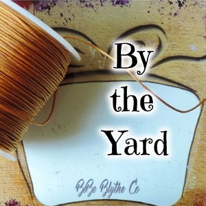 May include: A spool of golden-brown cord is featured, with the text "By the Yard" and the seller's name. The background has a painted, artistic design. The cord is likely for crafting or jewelry making.