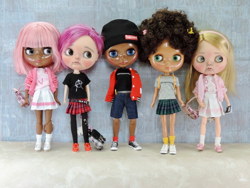 May include: Five dolls with different hairstyles and outfits. The dolls are wearing a pink sweater and skirt, a black t-shirt and plaid skirt, a red jacket and blue jeans, a grey t-shirt and plaid skirt, and a pink sweater and skirt. The dolls are all holding small bags.