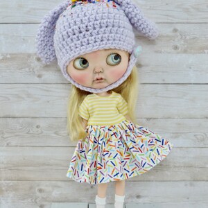 May include: A doll wearing a purple crocheted bunny hat with colorful sprinkles, a yellow and white striped shirt, and a white skirt with colorful sprinkles. The doll is standing on a light blue wooden surface.