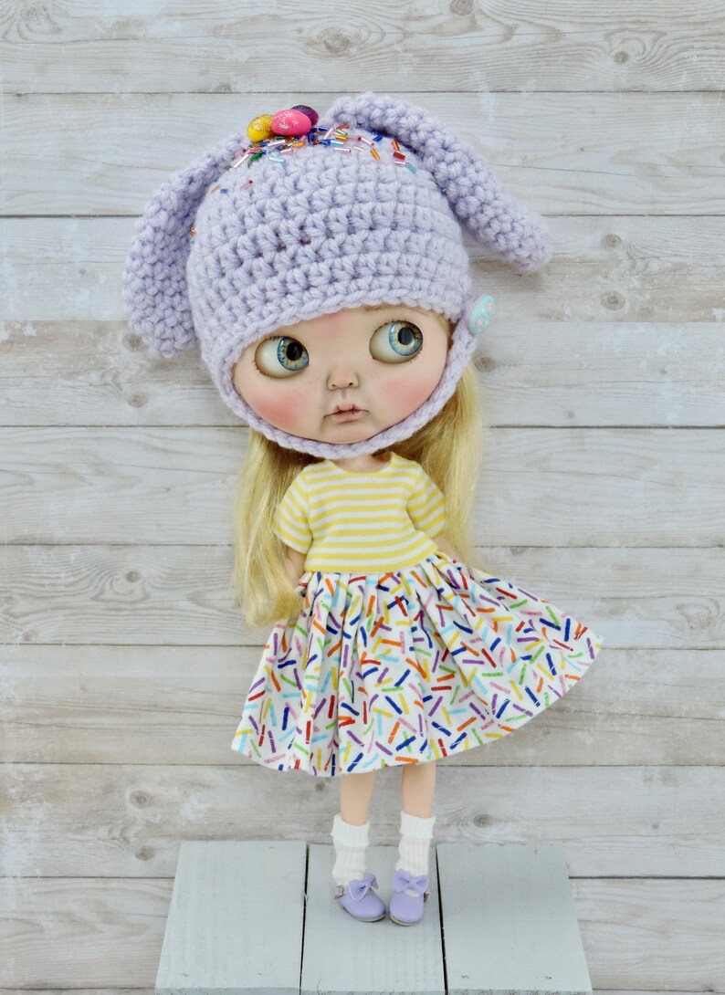 May include: A doll wearing a purple crocheted bunny hat with colorful sprinkles, a yellow and white striped shirt, and a white skirt with colorful sprinkles. The doll is standing on a light blue wooden surface.