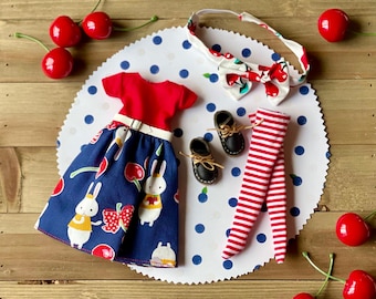 Könnte beinhalten: Ein Puppen-Outfit-Set mit einem rot-blauen Kleid mit Kirsch- und Hasenprint, einer rot-weiß gestreiften Leggings und einem Stirnband mit Schleife. Das Outfit befindet sich auf einem weißen Hintergrund mit blauen Punkten.