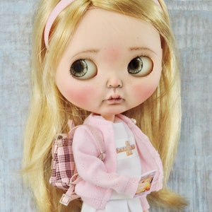 May include: A doll with blonde hair wearing a pink cardigan, a white shirt with the text "The Babysitter's Club" on it, a white skirt with pink trim, and a pink and white plaid backpack. The doll is holding a book.