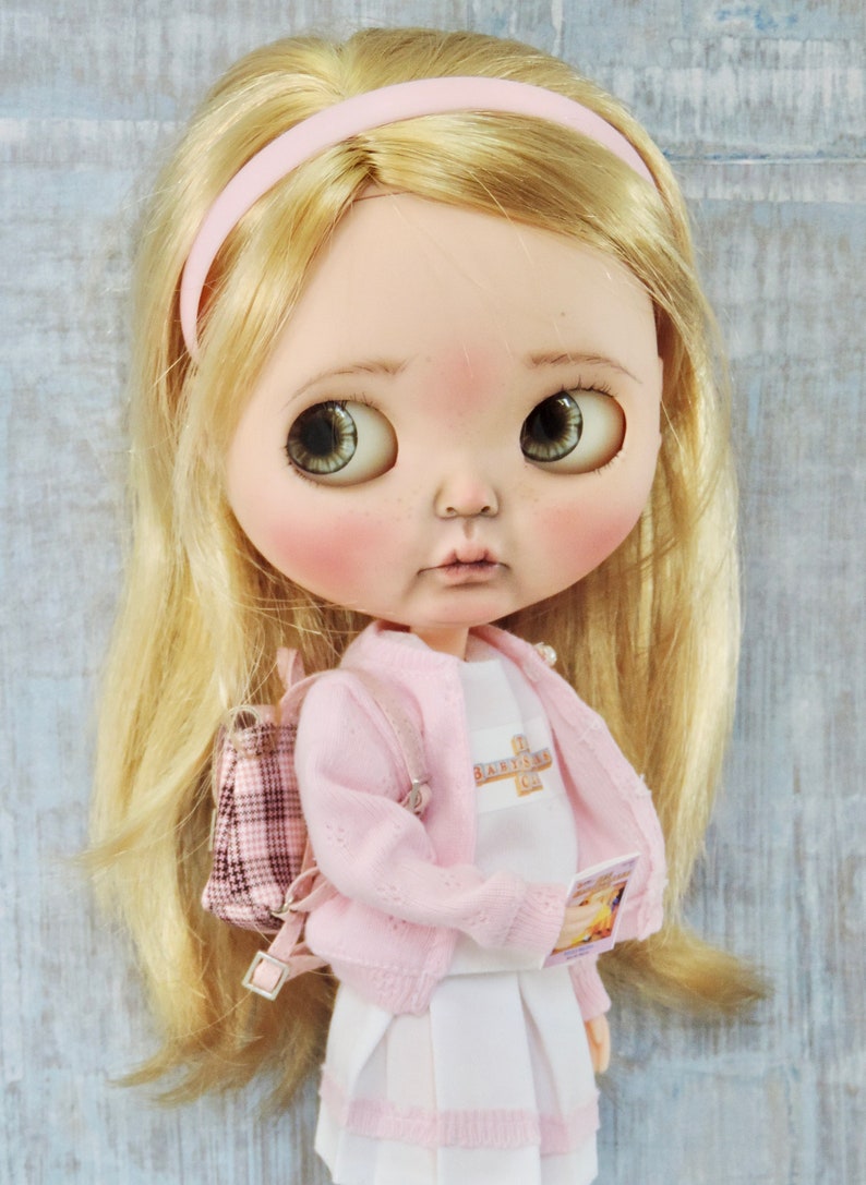 May include: A doll with blonde hair wearing a pink cardigan, a white shirt with the text "The Babysitter's Club" on it, a white skirt with pink trim, and a pink and white plaid backpack. The doll is holding a book.