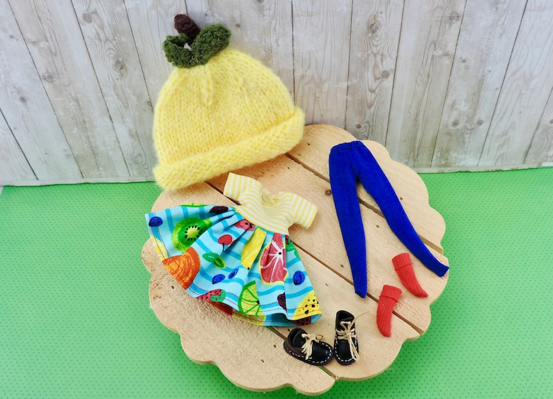 May include: A set of doll clothes including a yellow knitted hat with a green leaf detail, a blue and white dress with a fruit print, blue leggings, and red socks.
