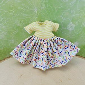 May include: A yellow and white striped short-sleeved shirt with a white skirt featuring a colorful confetti print. The skirt has a gathered waist and a full, flowing silhouette.