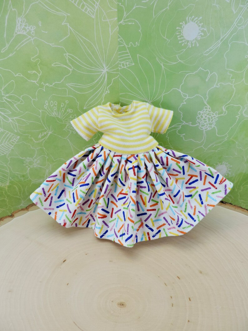 May include: A yellow and white striped short-sleeved shirt with a white skirt featuring a colorful confetti print. The skirt has a gathered waist and a full, flowing silhouette.